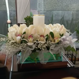 Arreglo floral mediano con orquídeas blancas, flores pequeñas, follaje y un cirio blanco en una base de cristal.