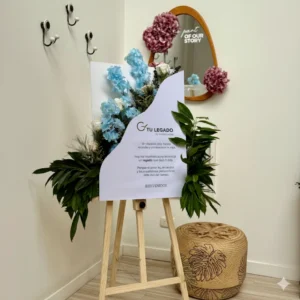 Caballete de madera con letrero "Tu Legado" adornado con flores azules y follaje verde, para eventos conmemorativos.