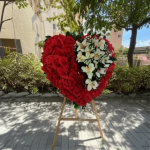 Arreglo floral fúnebre en forma de corazón grande, hecho con rosas rojas, apliques de orquídeas blancas y baby's breath sobre un trípode.
