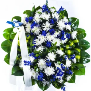 Corona fúnebre de tamaño mediano con flores de crisantemo blancas y amarillas, estatice, y hojas de caucho, sobre un soporte de madera.